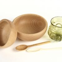Two carved wooden bowls with a wooden and a bone spoon, and a green glass beaker
